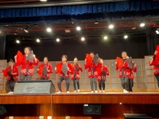 Unos 10 participantes interpretando una danza okinawense en el escenario, todos vestidos con trajes típicos japoneses “happi” 