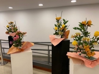 Tres arreglos de ikebana con flores amarillas y rosas, expuestas sobre un soporte en blanco y negro en el pasillo de entrada a la ceremonia de clausura
