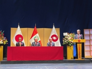 Embajador Yamamoto dirige sus palabras al lado de la mesa donde están el presidente saliente y el nuevo presidente en el escenario