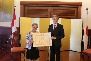 Consejera Igarashi con su diploma en mano y el Embajador delante del biombo y las banderas del Japón y del Perú