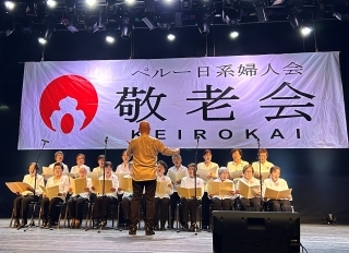 指揮者と18人ぐらいの婦人会会員によるコーラスの様子。