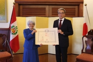Consejera Saito con su diploma en mano y el Embajador ubicados delante del biombo y las banderas del Japón y del Perú