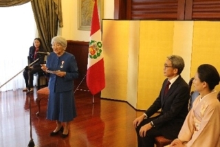 Embajador YAMAMOTO y su esposa sentados, escuchando las palabras de la consejera Saito que se encuentra de pie.