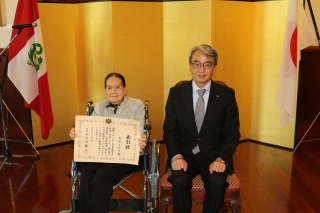 Señora TENYA con su diploma en mano y el Embajador ubicados delante del biombo y las banderas del Japón y del Perú.