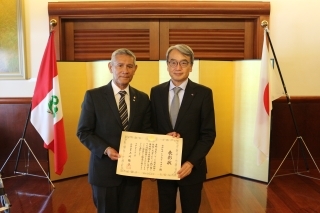 Señor Tokumori con su diploma en mano y el Embajador ubicados delante del biombo y las banderas del Japón y del Perú.