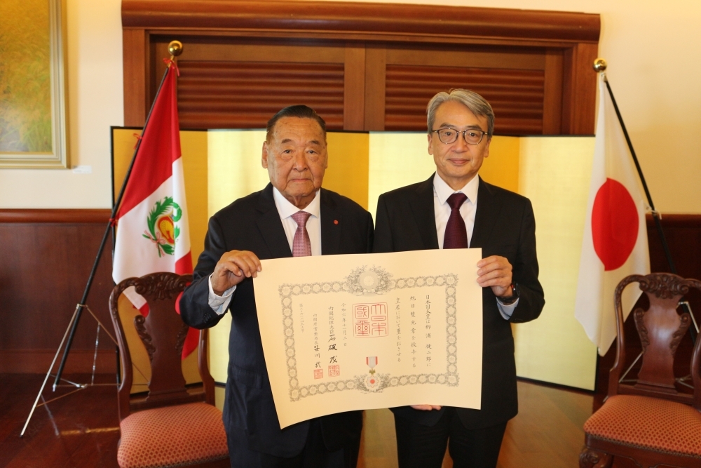 Consejero Yanaura con su diploma en mano y el Embajador ubicados delante del biombo y las banderas del Japón y del Perú