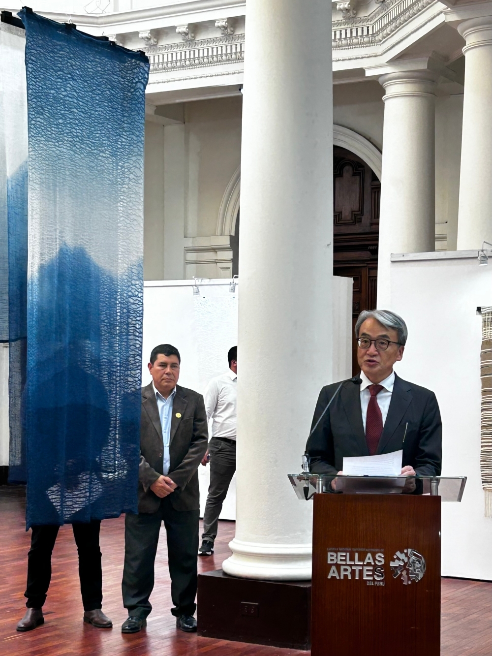 El Embajador YAMAMOTO brindando unas palabras durante la inauguración. Se puede apreciar una de las obras en tonos azulados del artista a la izquierda