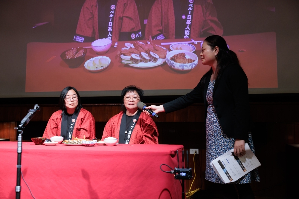A la izquierda, personales de la Asociación Peruano Japonesa realizando la demostración del uso de los palillos japoneses durante la conferencia. A la derecha la funcionaria SEINO Kanae sosteniendo el micrófono.
