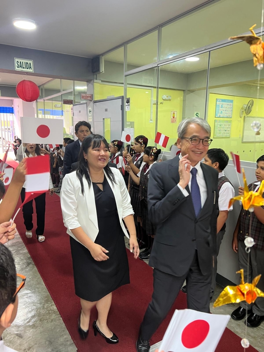 山元大使がオヤタ校長と生徒の歓迎を受けて学校の廊下を歩く様子