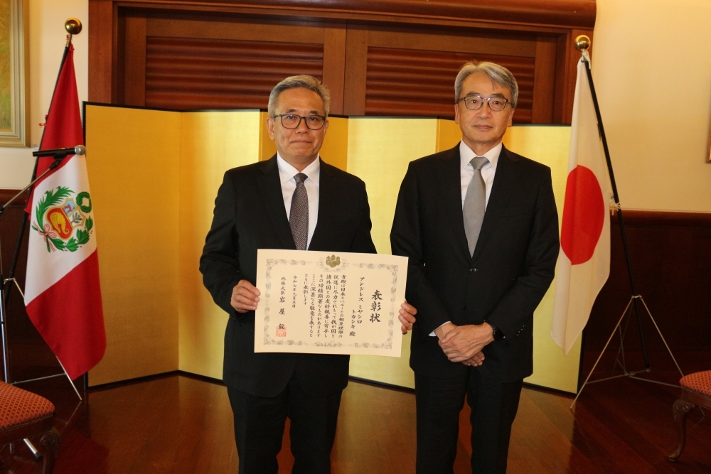El señor Miyashiro con su diploma en mano y el Embajador, ubicados delante del biombo y las banderas del Japón y del Perú