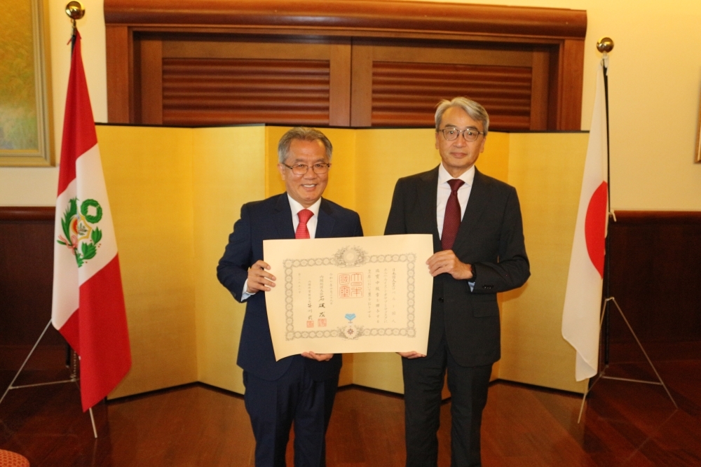 金屏風と両国国旗の前で、勲記を手に立つトオヤマ弁護士と山元大使