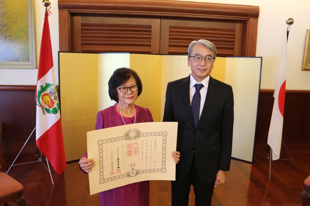 La señora Sakata con su diploma en mano y el embajador ubicados delante del biombo y las banderas del Japón y del Perú